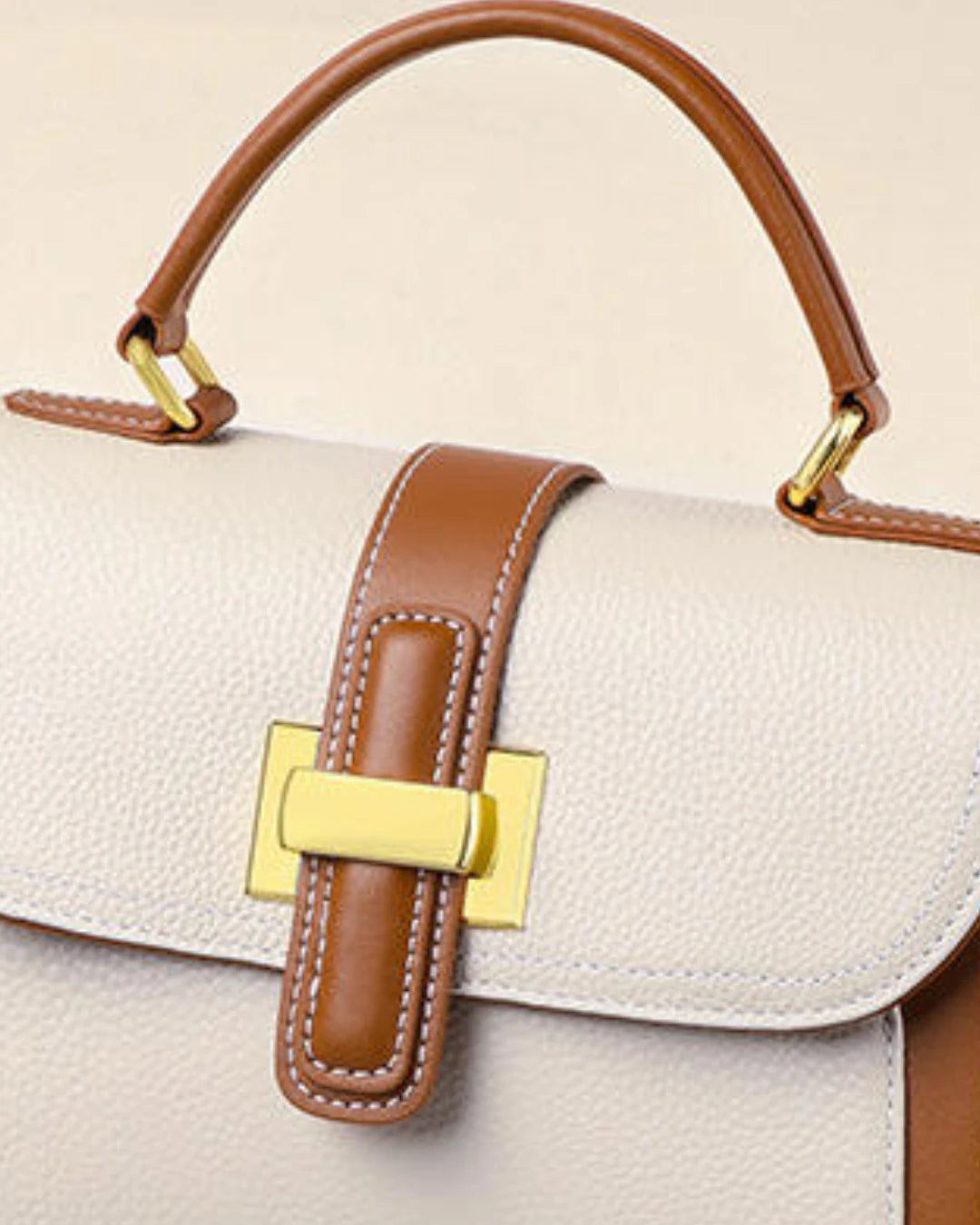 Detalhe frontal da bolsa feminina Maelle em couro bege, com tira caramelo, fecho metálico dourado, costuras claras e alça de mão.