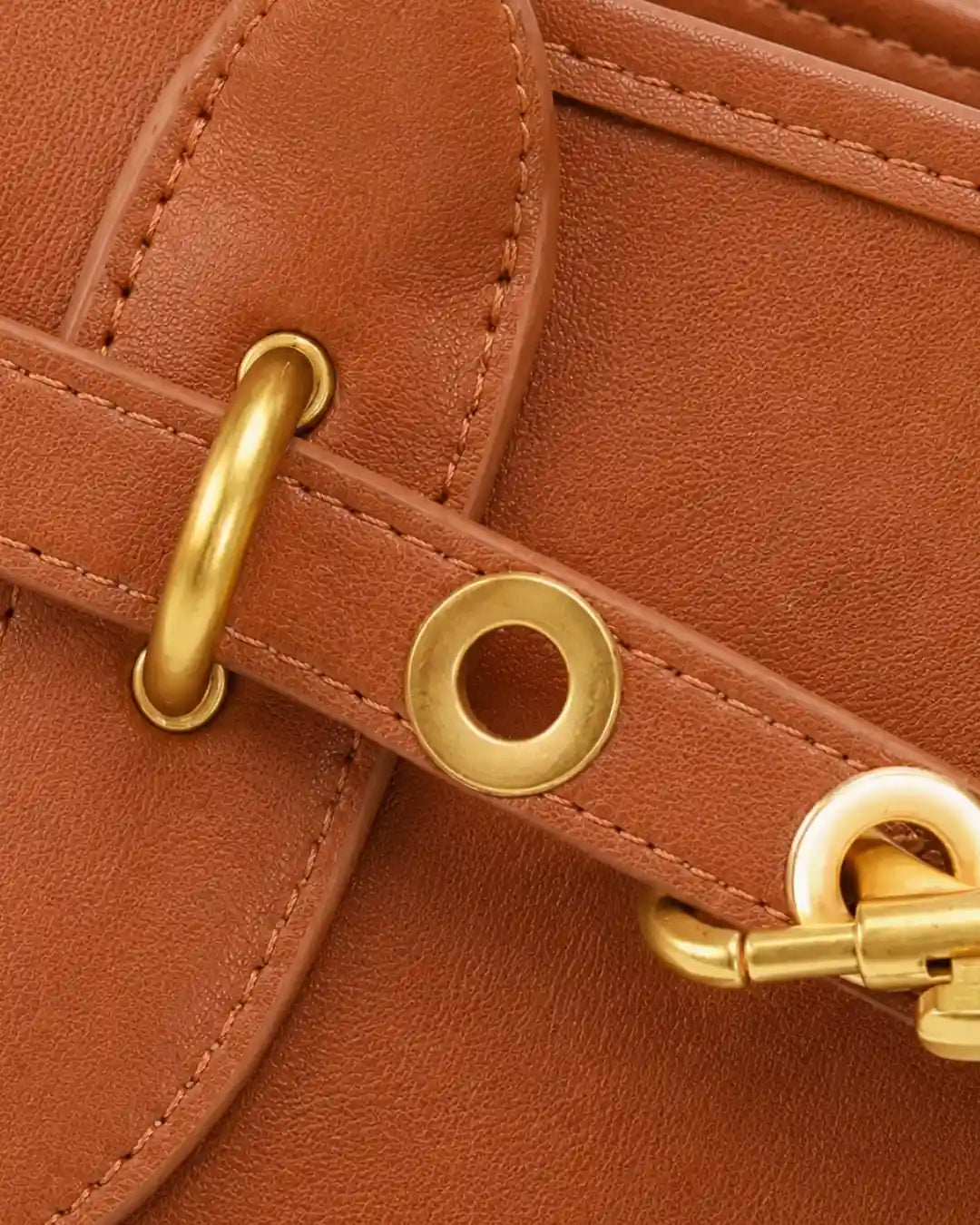 Detalhe frontal da bolsa feminina Aveline em couro caramelo, com tira decorativa, ilhós, argola e mosquetão dourado.