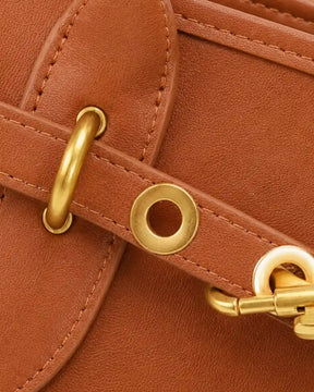 Detalhe frontal da bolsa feminina Aveline em couro caramelo, com tira decorativa, ilhós, argola e mosquetão dourado.