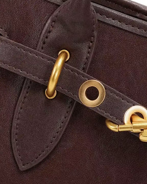 Detalhe frontal da bolsa feminina Aveline em couro marrom, com tira decorativa, ilhoses, argolas, mosquetão dourado e costuras aparentes.