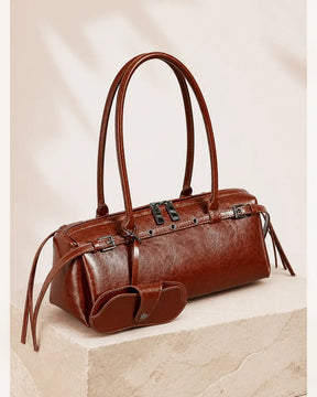 Bolsa feminina de couro marrom Celinne posicionada sobre uma base clara, com alças estruturadas, detalhes metálicos e porta-óculos combinando preso à frente.
