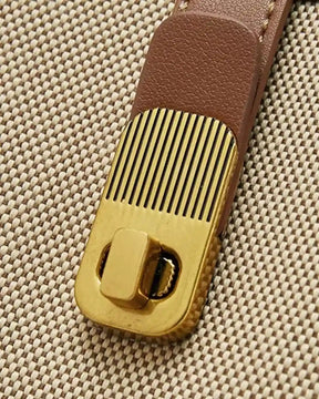 Detalhe do fecho giratório dourado da bolsa feminina de ombro bege Lisette, aplicado sobre tira marrom e corpo bege de textura tramada.