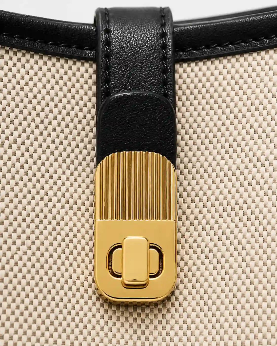 Detalhe do fecho giratório dourado da bolsa feminina de ombro preta Lisette, aplicado sobre tira preta e corpo bege com textura tramada.
