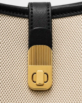 Detalhe do fecho giratório dourado da bolsa feminina de ombro preta Lisette, aplicado sobre tira preta e corpo bege com textura tramada.
