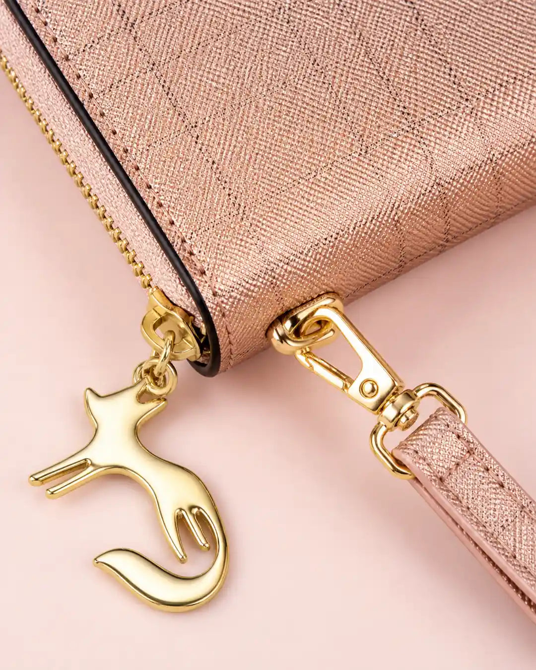 Detalhe da carteira feminina de couro rose Aurielle, destacando a textura do material, o fechamento em zíper dourado, a alça de pulso e o pingente metálico em formato de gato.

