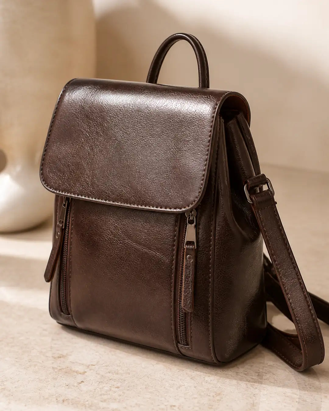 Mochila feminina de couro café Corinne em estúdio, com acabamento liso, aba frontal e alças ajustáveis.