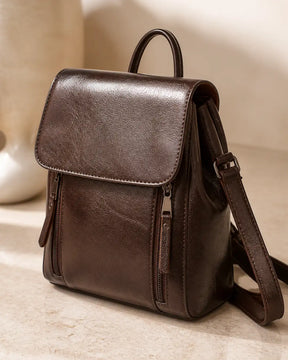 Mochila feminina de couro café Corinne em estúdio, com acabamento liso, aba frontal e alças ajustáveis.