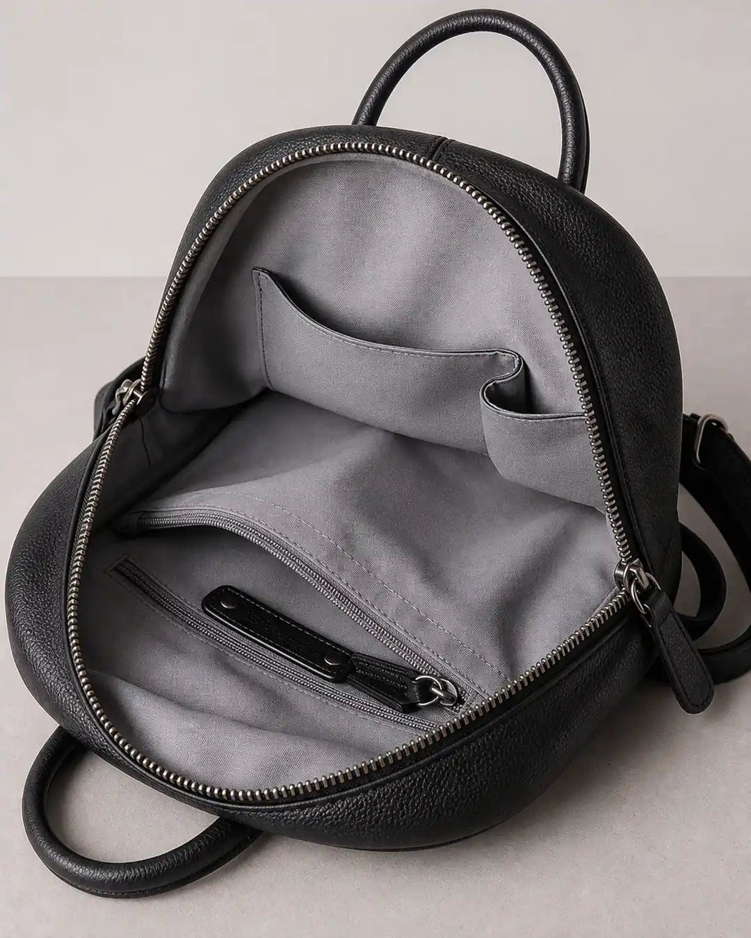 Mochila feminina de couro preta Aveline aberta, mostrando o interior com forro cinza, bolso interno com zíper e compartimentos organizadores.
